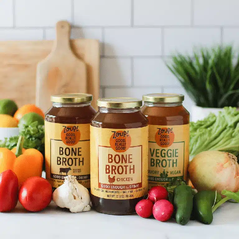 A collection of zoup broth jars surrounded by fresh vegetables on a marble countertop.