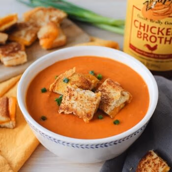 A white bowl filled with tomato soup topped with grilled cheese croutons and chives, in front of a jar of Zoup Chicken Broth