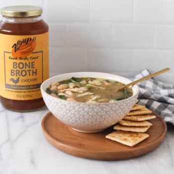 chicken soup in a white bowl served with crackers in front of a Zoup chicken bone broth jar on a marble counter.