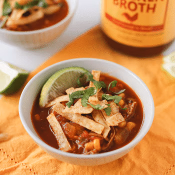 A white bowl filled with red soup and topped with crispy tortilla strips, cilantro and served with a lime wedge.