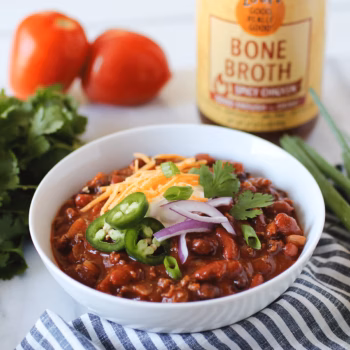 A shallow white bowl filled with heart chili, topped with sliced red onions, jalapenos, green onions, shredded cheese, cilantro and a dollop of sour cream.