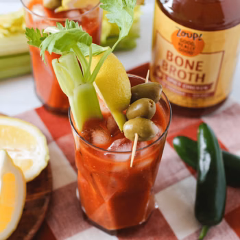 Spicy Bloody Mary garnished with celery, lemon wedge, and olives, served over ice with Zoup Spicy Chicken bone broth, jalapeños, and lemon slices in the background.