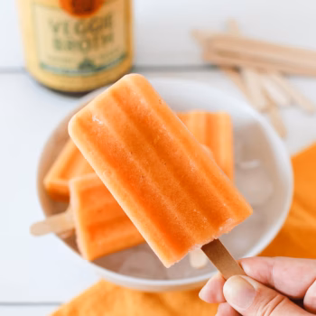 Closeup of a person's hand holding a frozen mango popsicle, in front of a tray of popsicles and a jar of Zoup Veggie Broth.