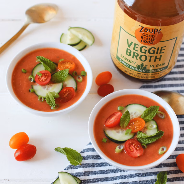 Two white bowls filled with blended tomato gazpacho topped with sliced cucumber, cherry tomatoes, green onion and sprigs of mint, beside a jar of Zoup Veggie Broth