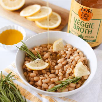 A white bowl filled with brothy white beans and topped with rosemary and lemon slices, in front of a jar of Zoup Veggie Broth.