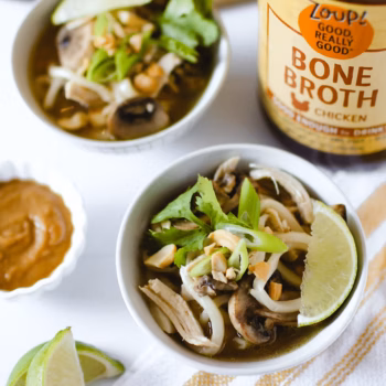 Bowl of chicken udon soup with mushrooms, noodles, lime wedge, chopped peanuts, green onions, and cilantro, set beside a jar of Zoup chicken bone broth and a small dish of peanut sauce.