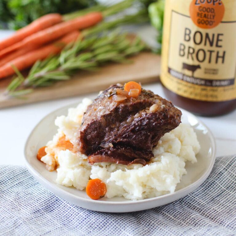 Beef short rib topped with carrots and onions, served over mashed potatoes on a plate, with a jar of Zoup Beef bone broth and fresh herbs in the background.