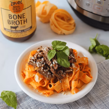 Plate of wide pappardelle pasta topped with shredded beef ragu and fresh basil, surrounded by dry pasta nests, a jar of bone broth, and an Instant Pot in the background.
