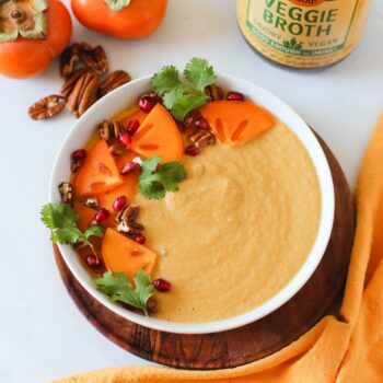 A white bowl filled with creamy soup topped with pomegranate seeds, sliced persimmons and pecan pieces, in front of a jar of Zoup Veggie Broth.
