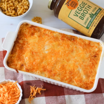 A white casserole dish filled with baked macaroni and cheese, topped with crispy melted cheese, beside a jar of Zoup Veggie Broth.