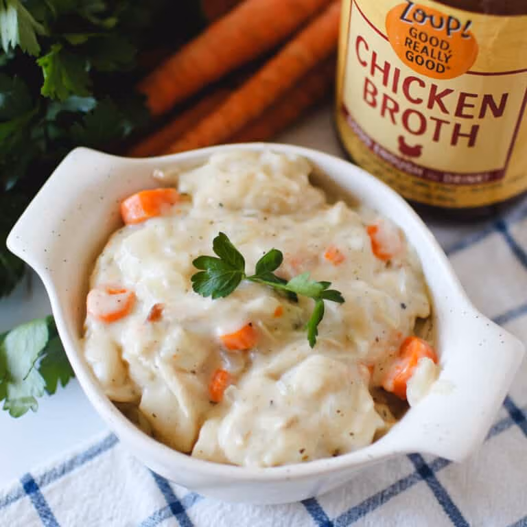 A white bowl filled with creamy soup with carrots, chicken, onions and dumplings and topped with a sprig of parsley, served beside a jar of Zoup Chicken Broth