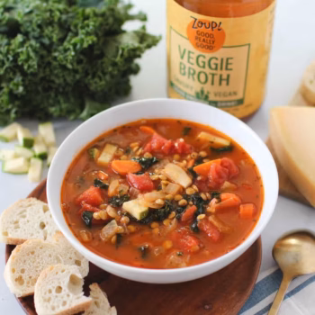 Hearty vegetable soup in a white bowl, filled with lentils, tomatoes, carrots, zucchini, kale, and onion in a rich tomato broth, served with slices of crusty bread and a bottle of Zoup veggie broth in the background.