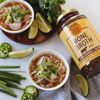 A white bowl filled with rice noodles in broth, garnished with sliced jalapeño, green onion, and cilantro, with a jar of Zoup beef bone broth in the background.