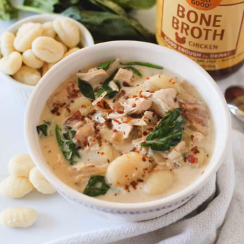 Creamy chicken gnocchi soup in a white bowl, filled with tender gnocchi, shredded chicken, spinach, and crumbled bacon, with a jar of Zoup chicken bone broth in the background.