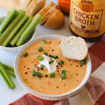 A bowl of creamy zoup topped with chopped green onions, sour cream and served with a slice of bread, placed in front of a jar of Zoup Chicken Broth.