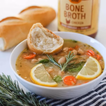 White bean soup with carrots, garnished with a rosemary sprig and lemon slices. A baguette and a bottle of Zoup chicken bone broth are visible in the background.
