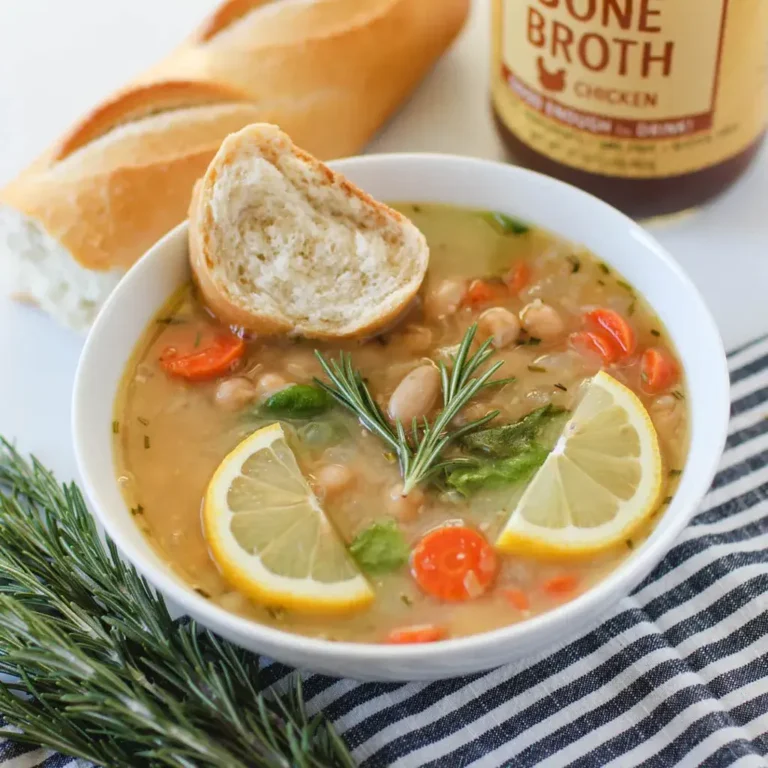 White bean soup with carrots, garnished with a rosemary sprig and lemon slices. A baguette and a bottle of Zoup chicken bone broth are visible in the background.