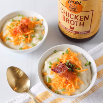 Two bowls of creamy potato soup and topped with diced bacon, chives, and shredded cheese, beside a jar of Zoup Chicken Broth
