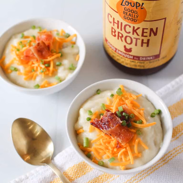 Two bowls of creamy potato soup and topped with diced bacon, chives, and shredded cheese, beside a jar of Zoup Chicken Broth