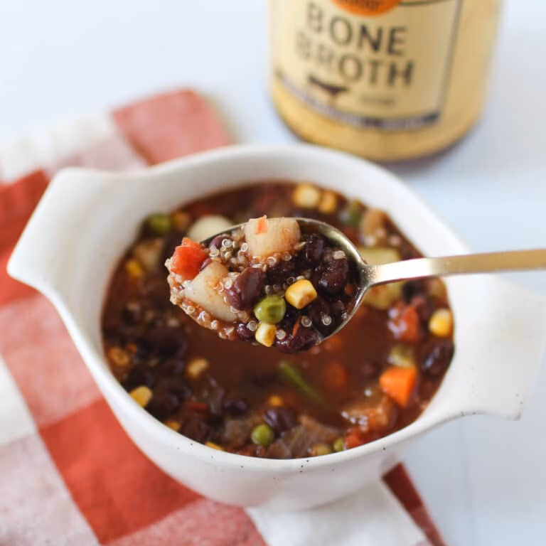 Closeup of a spoon filled with vegetable soup containing potatoes, peas, corn, carrots, beans, and quinoa, in front of a bowl filled with the same soup.