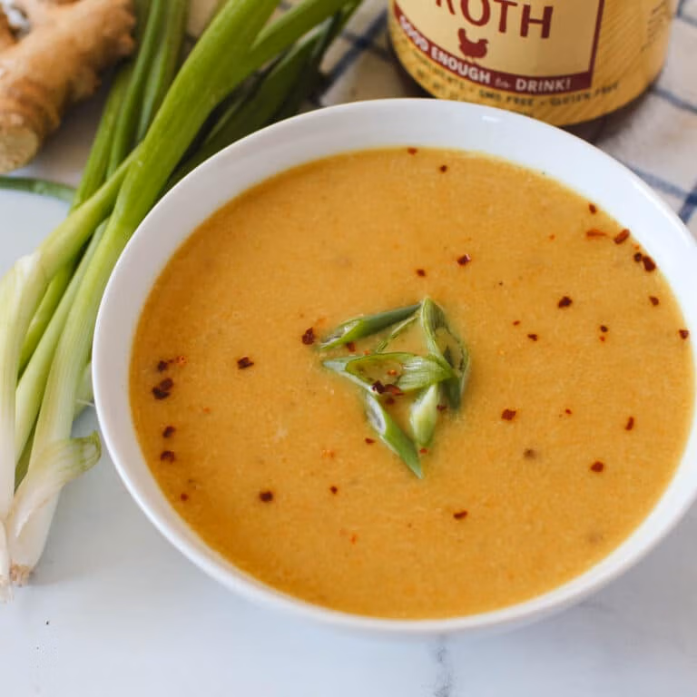 Blended butternut squash soup in a white bowl topped with green onions and red pepper flakes in front of a jar of Zoup Chicken Bone Broth