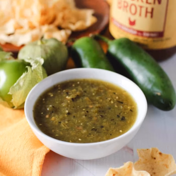 A white bowl filled with salsa verde surrounded by tomatillos, jalapeno peppers, tortilla chips and a jar of Zoup Chicken Broth.