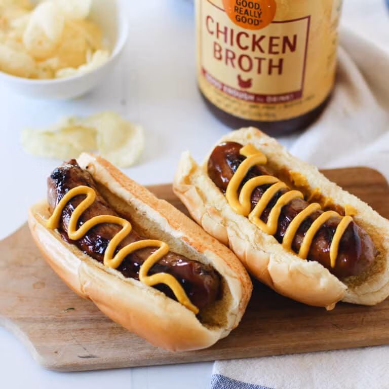 Two grilled sausages in buns topped with mustard, served on a wooden board with a bowl of potato chips and a jar of Zoup chicken broth in the background.