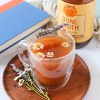 A clear mug filled with broth and topped with dasies and a lavender sprig in front of a jar of Zoup Chicken Bone Broth