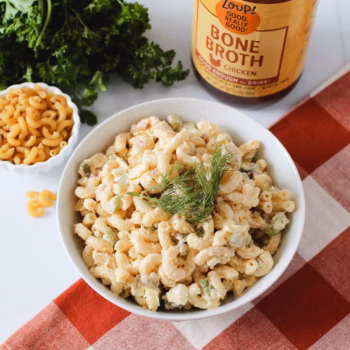 Creamy macaroni salad in a white bowl topped with a sprig of dill, placed on a checkered red napkin in front of a jar of Zoup Chicken Bone Broth