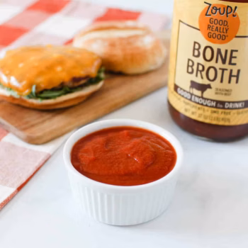A white ramekin filled with bright red ketchup in front of a deconstructed burger and a jar of Zoup Beef Bone Broth.