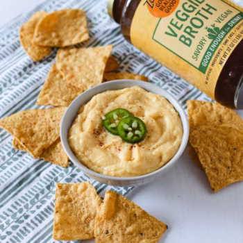 a white bowl filled with creamy dip topped with jalapenos, surrounded by scattered tortilla chips and a jar of Zoup Veggie Broth