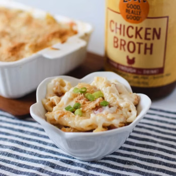 A white flower shaped bowl filled with creamy pasta and toppe with green onion, melted cheese, and breadcrumbs, in front of a jar of Zoup Chicken Broth