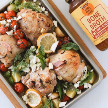 A sheet pan filled with roasted chicken thighes, potatoes, feta cheese, lemon slices, and cherry tomatoes, topped with mint and dill, beside a jar of Zoup Spicy Chicken Bone Broth