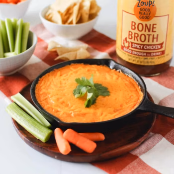 A mini skillet filled with creamy dip topped with melted cheese on a wooden plate surrounded by chopped veggies, in front of a jar of Zoup Spicy Chicken Bone Broth