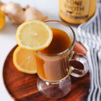 A clear mug filled with bone broth with a single lemon slice placed on the corner of the mug, and a jar of Zoup Beef Bone Broth behind it.
