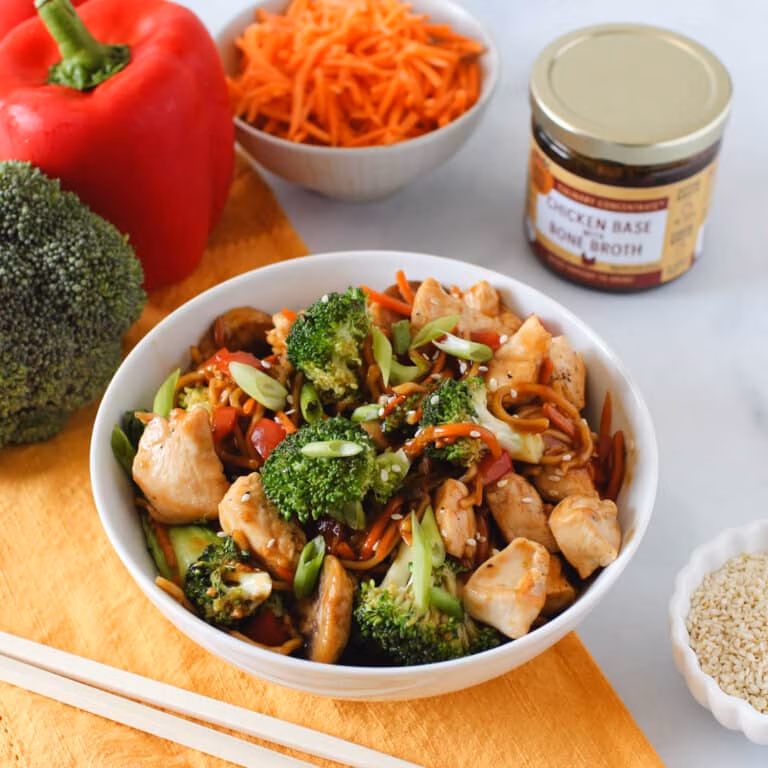 Bowl of chicken stir-fry with broccoli, red peppers, carrots, noodles, and green onions, surrounded by fresh vegetables, sesame seeds, and a jar of Zoup chicken broth concentrate.