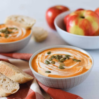 A white bowl filled with swirled creamy pumpkin soup topped with pumpkin seeds with sliced bread beside it on a red checked napkin.