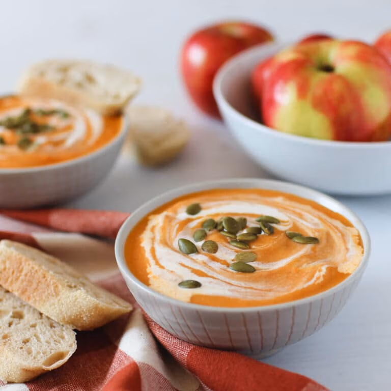 A white bowl filled with swirled creamy pumpkin soup topped with pumpkin seeds with sliced bread beside it on a red checked napkin.