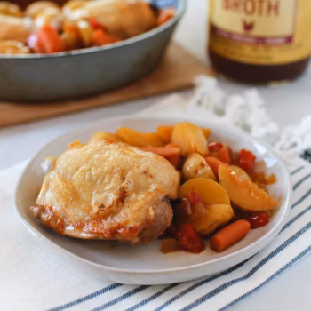 Plate of roasted chicken thigh with potatoes, carrots, and red bell peppers in broth, set in front of a skillet and a jar of chicken bone broth.