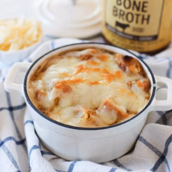 Mini white Dutch oven filled with French onion soup, topped with golden melted cheese and bread, set on a blue checkered towel with a jar of Zoup beef bone broth in the background