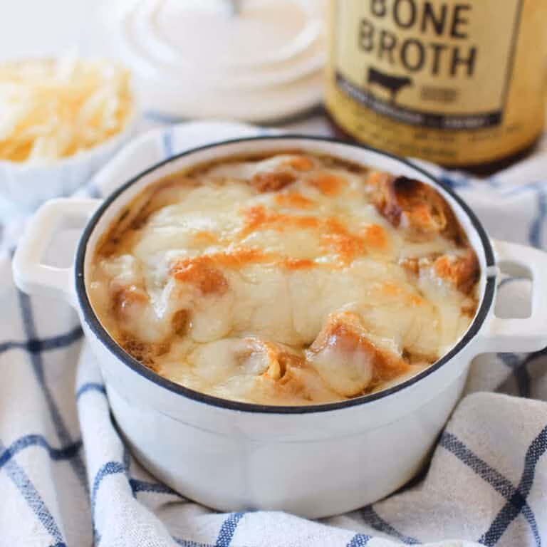 Mini white Dutch oven filled with French onion soup, topped with golden melted cheese and bread, set on a blue checkered towel with a jar of Zoup beef bone broth in the background
