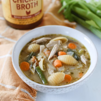 Bowl of chicken vegetable soup with shell pasta, shredded chicken, carrots, celery, and green beans in a savory broth, set beside a jar of Zoup chicken broth and fresh celery.