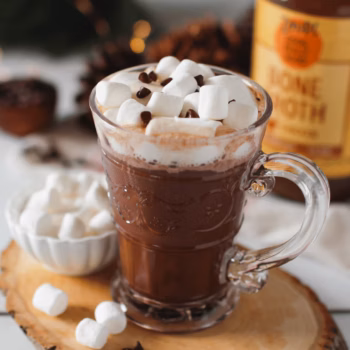 Glass mug of hot chocolate topped with mini marshmallows and chocolate chips, set on a wood slice with extra marshmallows and a jar of Zoup bone broth in the background.