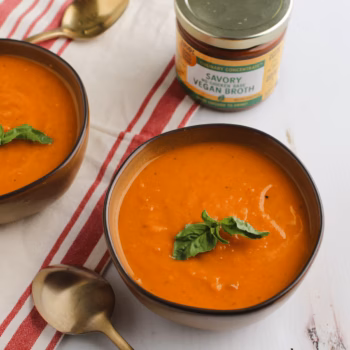 Blended pepper and sweet potato soup in two brown bowls next to a jar of Zoup Savory Vegan Broth Concentrate on a red striped towel