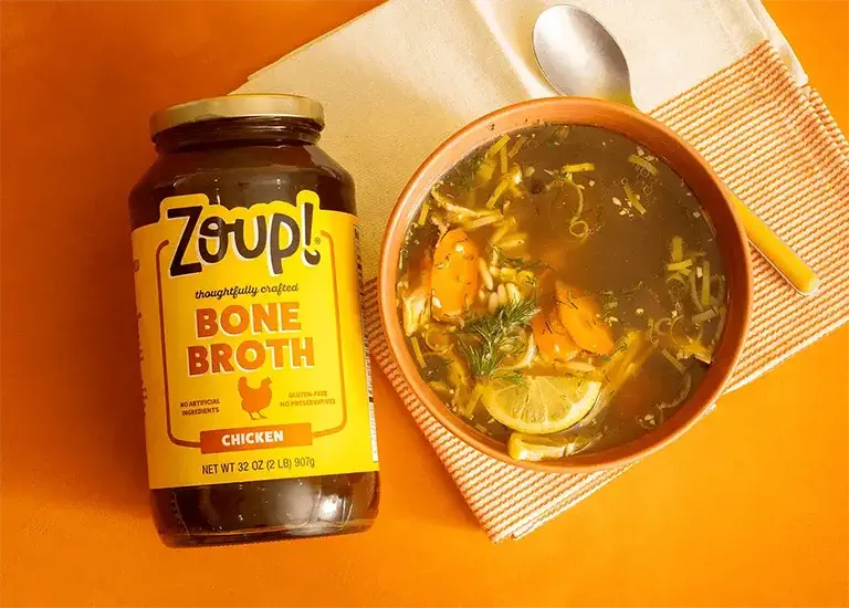 A jar of Zoup chicken bone broth lays flat on an orange table, beside a bowl of chicken soup