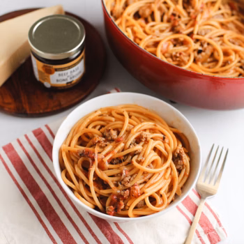 A bowl of bucatini pasta with ground sausage, beside a red dutch oven filled with the same pasta, and a jar of Zoup Beef Base Contentrate in the background.