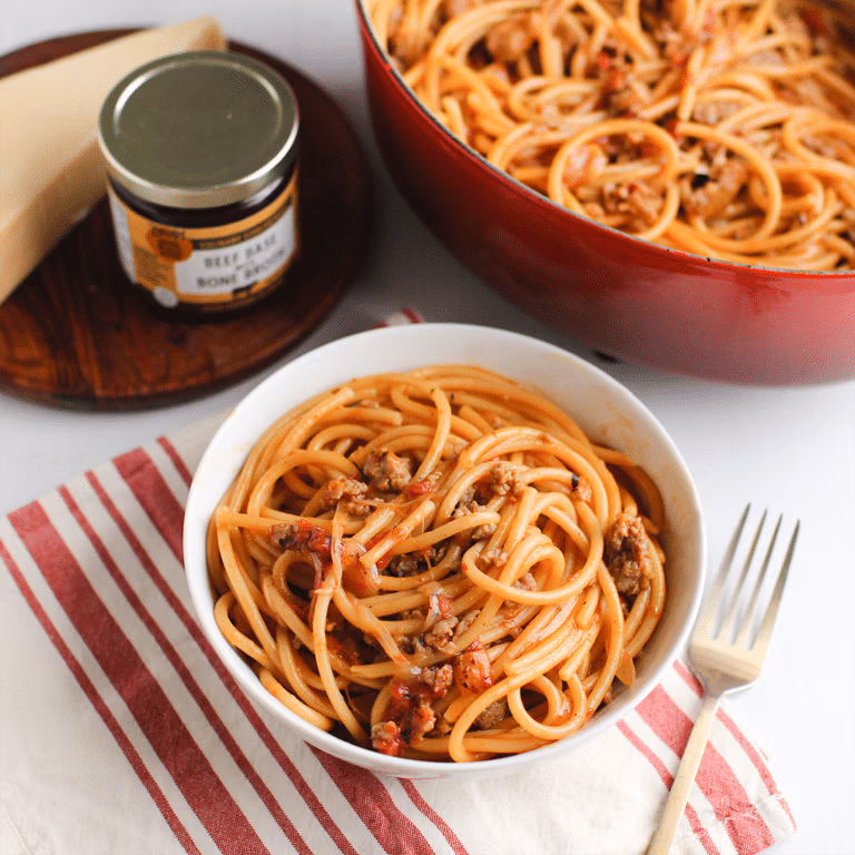 A bowl of bucatini pasta with ground sausage, beside a red dutch oven filled with the same pasta, and a jar of Zoup Beef Base Contentrate in the background.
