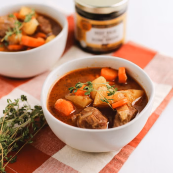 Two white bowls filled with brothy soup, diced carrots and beef, placed in front of a jar of Zoup Beef Base Concentrate