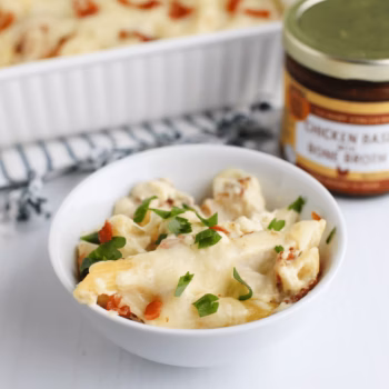 A white bowl filled with chicken and noodles topped with melted cheese and chopped parsley, with a jar of Zoup chicken base concentrate in the background.
