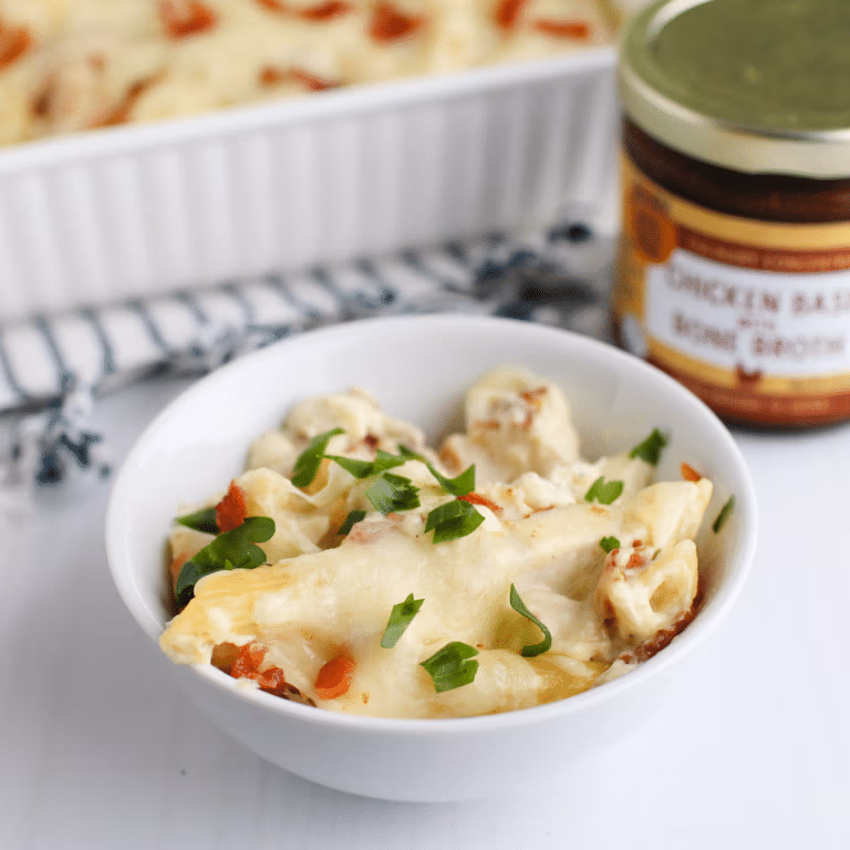 A white bowl filled with chicken and noodles topped with melted cheese and chopped parsley, with a jar of Zoup chicken base concentrate in the background.